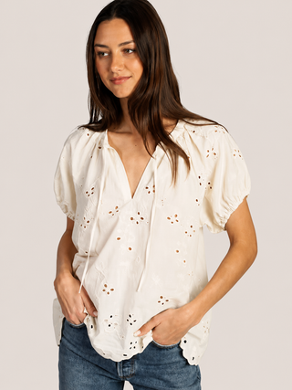 A woman wearing a white cream-colored eyelet blouse with puff sleeves and a split neckline, paired with blue jeans, against a solid light beige background.