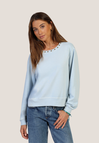 A woman modeling a light blue crewneck sweatshirt with silver metal stud detailing around the neckline, paired with blue jeans, against a solid light beige background.