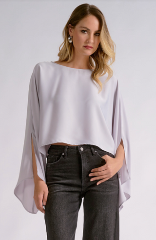A woman modeling a light lavender-purple draped blouse with wide, flowing sleeves and a boat neckline, paired with dark gray jeans, against a light gray studio background.