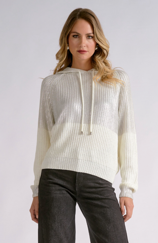 A woman modeling a textured, color-blocked knit hoodie in cream and white, paired with dark gray jeans, against a light gray studio background.