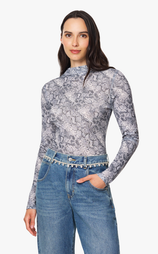 A woman modeling a long-sleeve mock neck top with a gray and white snakeskin pattern, paired with blue jeans and a beaded belt, against a plain white background.