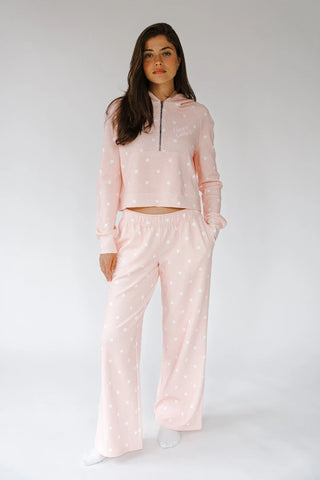 A woman modeling a light pink two-piece loungewear set with white polka dots, consisting of a cropped long-sleeve half-zip hoodie and matching wide-leg sweatpants, against a plain light gray studio background.