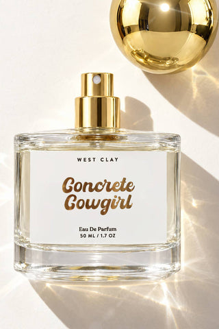 A 50 mL clear glass perfume bottle with a gold spray top and a white label reading 'WEST CLAY Concrete Cowgirl Eau De Parfum', photographed on a bright white background with dramatic sunlight and shadows. A large reflective gold sphere is partially visible in the upper right.