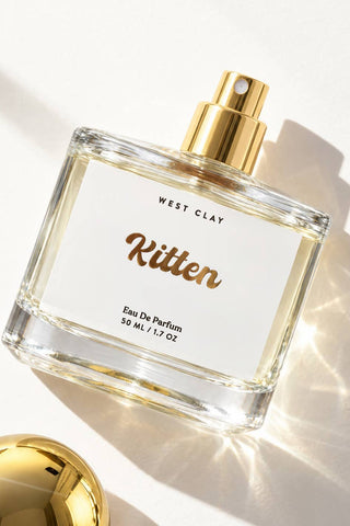 A clear rectangular perfume bottle with a white label reading 'WEST CLAY Kitten Eau De Parfum 50 ML / 1.7 OZ' and a shiny gold spray top, photographed on a bright white surface with strong sunlight and shadows.