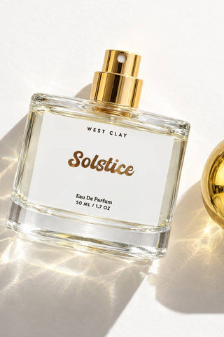 A clear glass perfume bottle labeled West Clay Solstice Eau De Parfum with a gold spray top, shown on a white background with a separate gold spherical cap beside it and bright sunlight casting shadows.