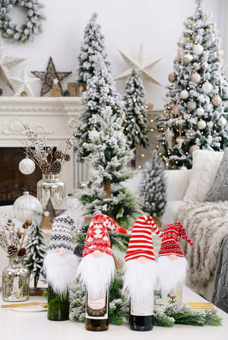 A set of wine bottle covers designed as festive gnomes with white beards and Christmas-themed hats, displayed against a holiday decor background.
