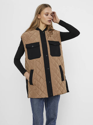 A female model wearing a beige and black long quilted waistcoat with chest pockets and front pockets, paired with a black top and blue jeans.