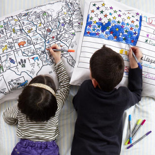 Two children lying on a bed coloring on white pillowcases with markers.